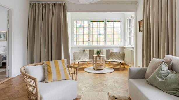 Una habitación con sofá, sillón y una mesita con una planta. Hay cortinas, una ventana con vistas y un radiador.