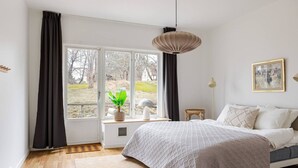 Un dormitorio con un ventanal grande, una cama, una lámpara colgante y una mesita de noche con lámpara.