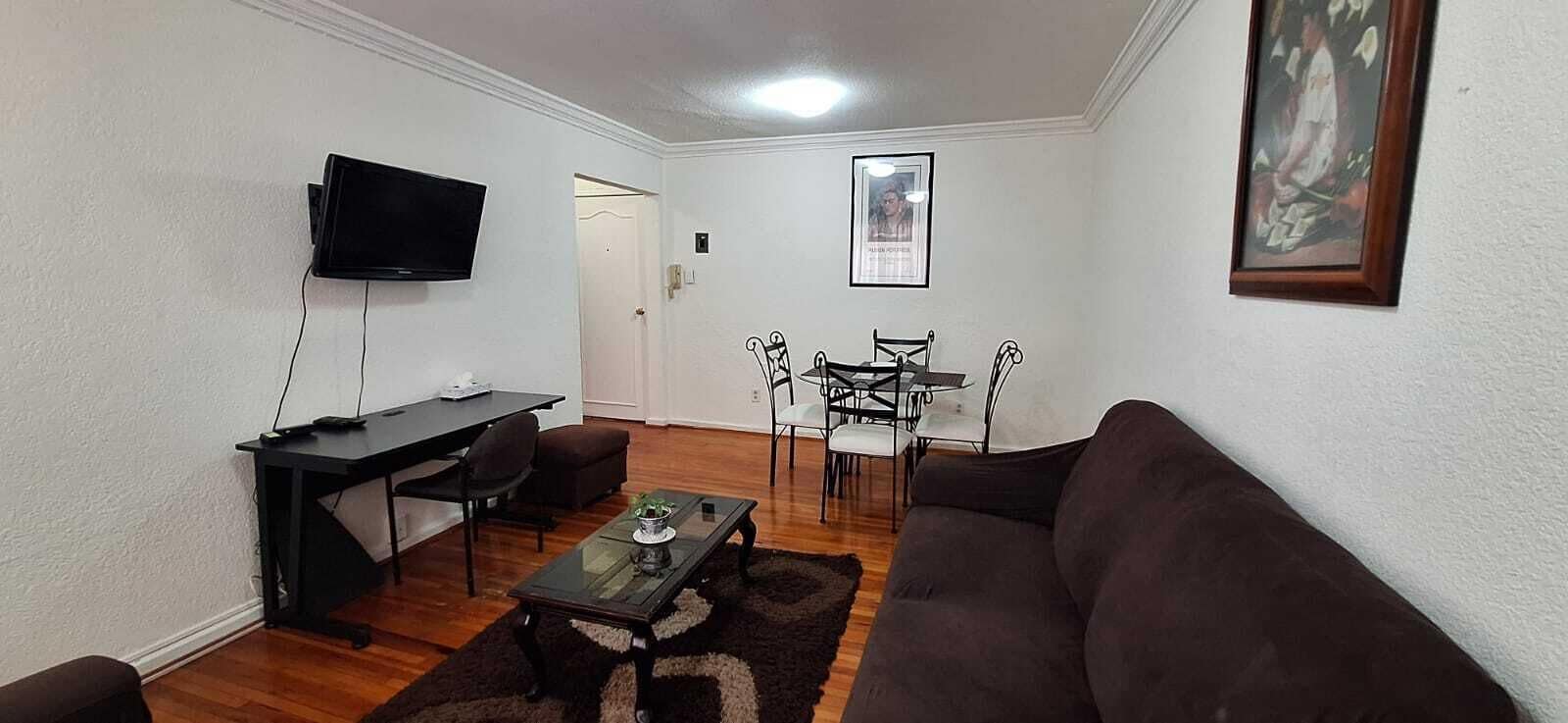 Un saló amb un sofà, una taula de centre, una televisió, una taula de menjador amb caires i un quadre al mur.