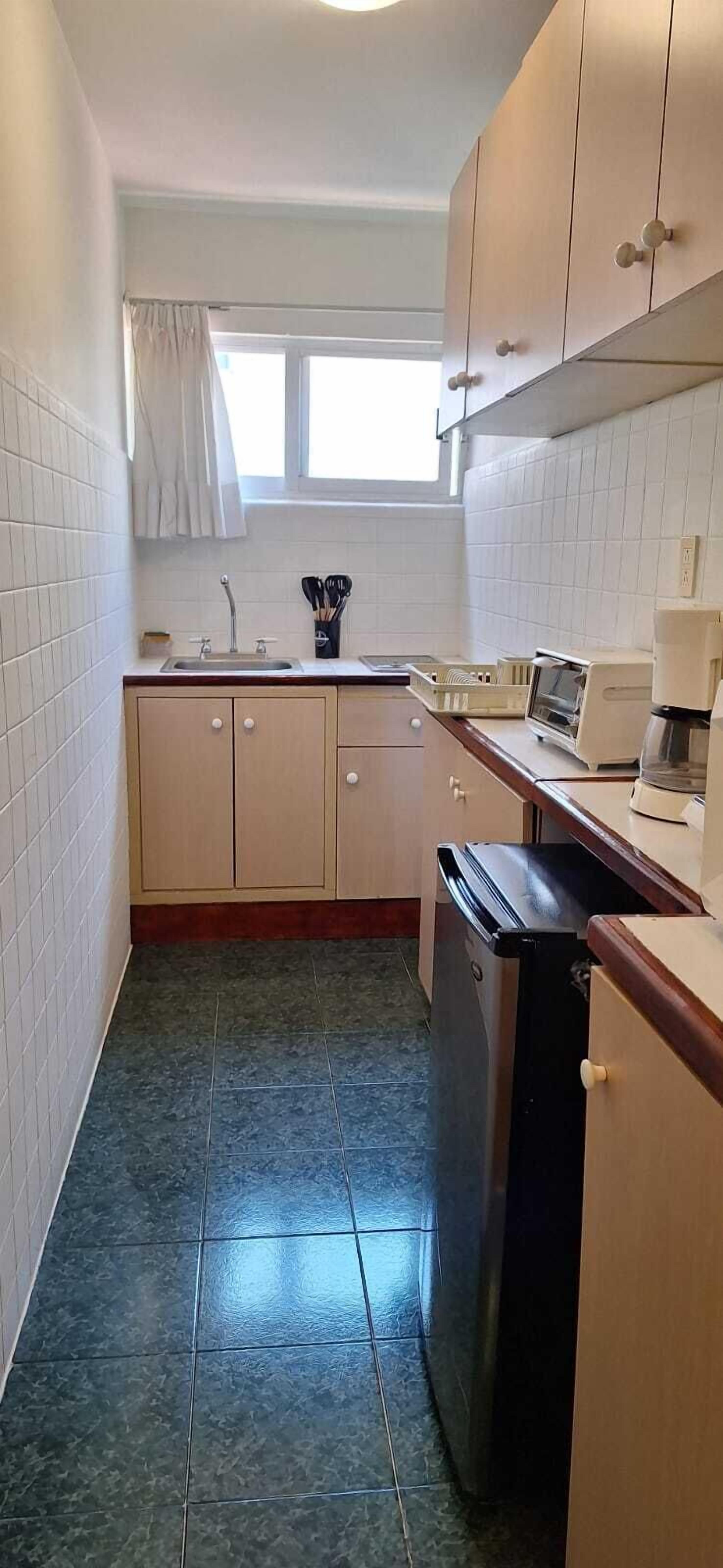 A narrow kitchen with white tiled walls, a window with white curtains, a sink, cabinets, a dishwasher, and a coffee maker.