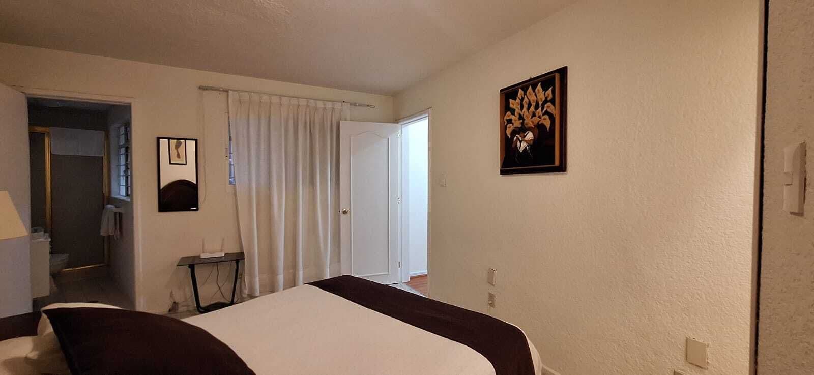 Une chambre d’hôtel avec un lit, une porte, une fenêtre avec des rideaux, un tableau accroché au mur et une petite table avec une lampe.