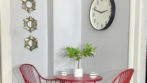 Een eetgedeelte met een ronde tafel, twee rode stoelen, een potplant en een wandklok.