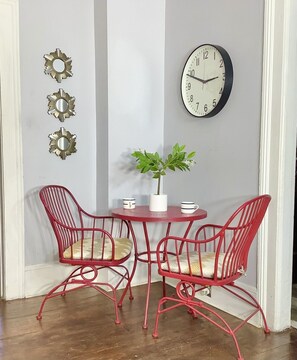 Una habitación con una mesa redonda, dos sillas rojas, una planta en maceta, un reloj de pared y tres espejos decorativos.