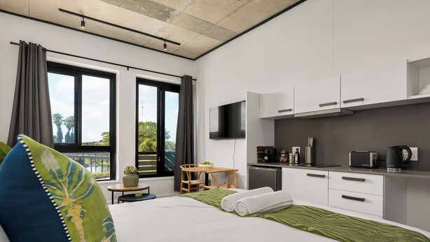 Et moderne hotelværelse med en seng, et lille bord, en stol, et tekøkken og store vinduer.