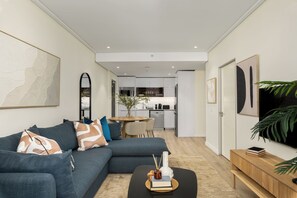 A modern living room with a sectional sofa, a coffee table with books and a vase, a dining area with a round table and chairs, and a kitchen in the background. - Luxurious Apartment With View Pool and Gym (Cape Town)