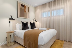 A neatly made bed with a brown blanket, two black pillows, and a white pillow. There are two bedside lamps, a round floor lamp, and a framed abstract painting on the wall. - Luxurious Apartment With View Pool and Gym (Cape Town)