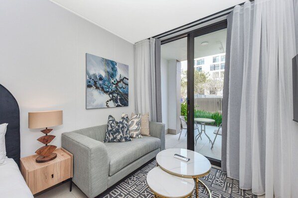Et moderne hotelværelse med en grå sofa, et rundt hvidt bord, et billede på væggen og en balkon med havemøbler.