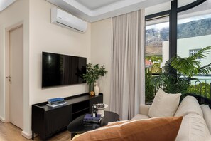 A modern living room with a flat-screen TV, a black coffee table, a sofa, a potted plant, and a view of a mountainous landscape through large windows. - Designer Apartment in Vredehoek | Mountain Views (Cape Town)