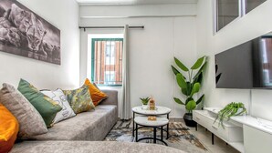 A modern living room with a grey sofa, a small round table, a TV mounted on the wall, and a large window with a view of a brick building. - Chic 2BR in Illovo: Work, Play, Unwind (Johannesburg)