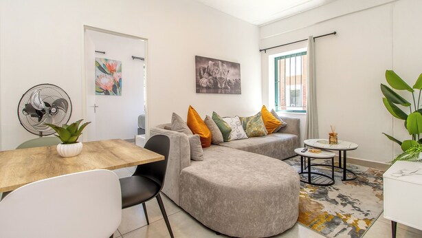 A modern living room with a sofa, a dining table, a fan, and a window with curtains. - Chic 2BR in Illovo: Work, Play, Unwind (Johannesburg)