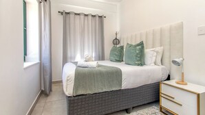 A bedroom with a bed, a bedside table, a lamp, and a window with curtains. - Chic 2BR in Illovo: Work, Play, Unwind (Johannesburg)