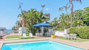 A swimming pool with lounge chairs and a table, surrounded by palm trees and a building with a blue roof.