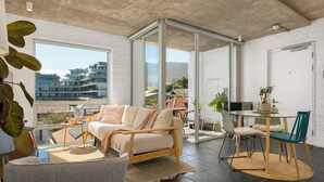 A modern living room with a sofa, a dining table with chairs, a large window, and a view of buildings. - Sunny Sea Point Stay with Balcony and Views (Cape Town)