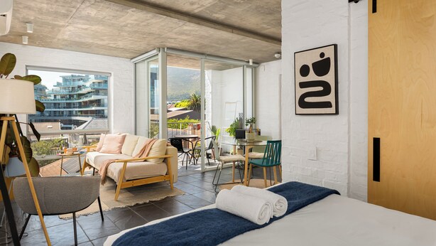 A modern hotel room with a bed, a sofa, a desk, and a balcony with a view of buildings. - Sunny Sea Point Stay with Balcony and Views (Cape Town)