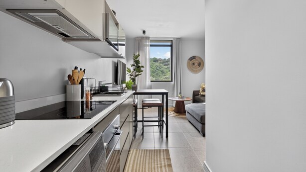 A modern kitchen with a central island, a dining area with a round table, and a view of greenery outside. - Stylish 2 Bedroom | Zimbali | Pool and Braai (Dolphin Coast)
