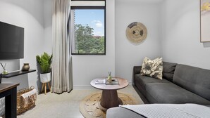 A modern living room with a grey sofa, a round coffee table, a TV on the wall, a window with curtains, and a decorative wall piece. - Stylish 2 Bedroom | Zimbali | Pool and Braai (Dolphin Coast)