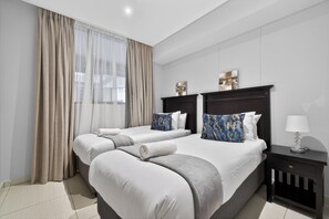 A hotel room with two beds, a nightstand, a lamp, and a window with curtains. - Palm Boulevard 404 | 3Bed Luxury - Gateway (Umhlanga)