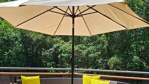 Um terraço com uma mesa redonda, um guarda-sol bege e móveis de vime com almofadas amarelas.