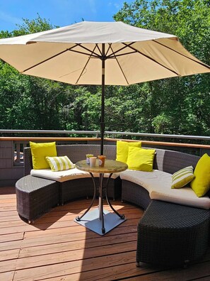 Una terraza con una mesa redonda, un paraguas color beige y mobiliario de mimbre con cojines amarillos.
