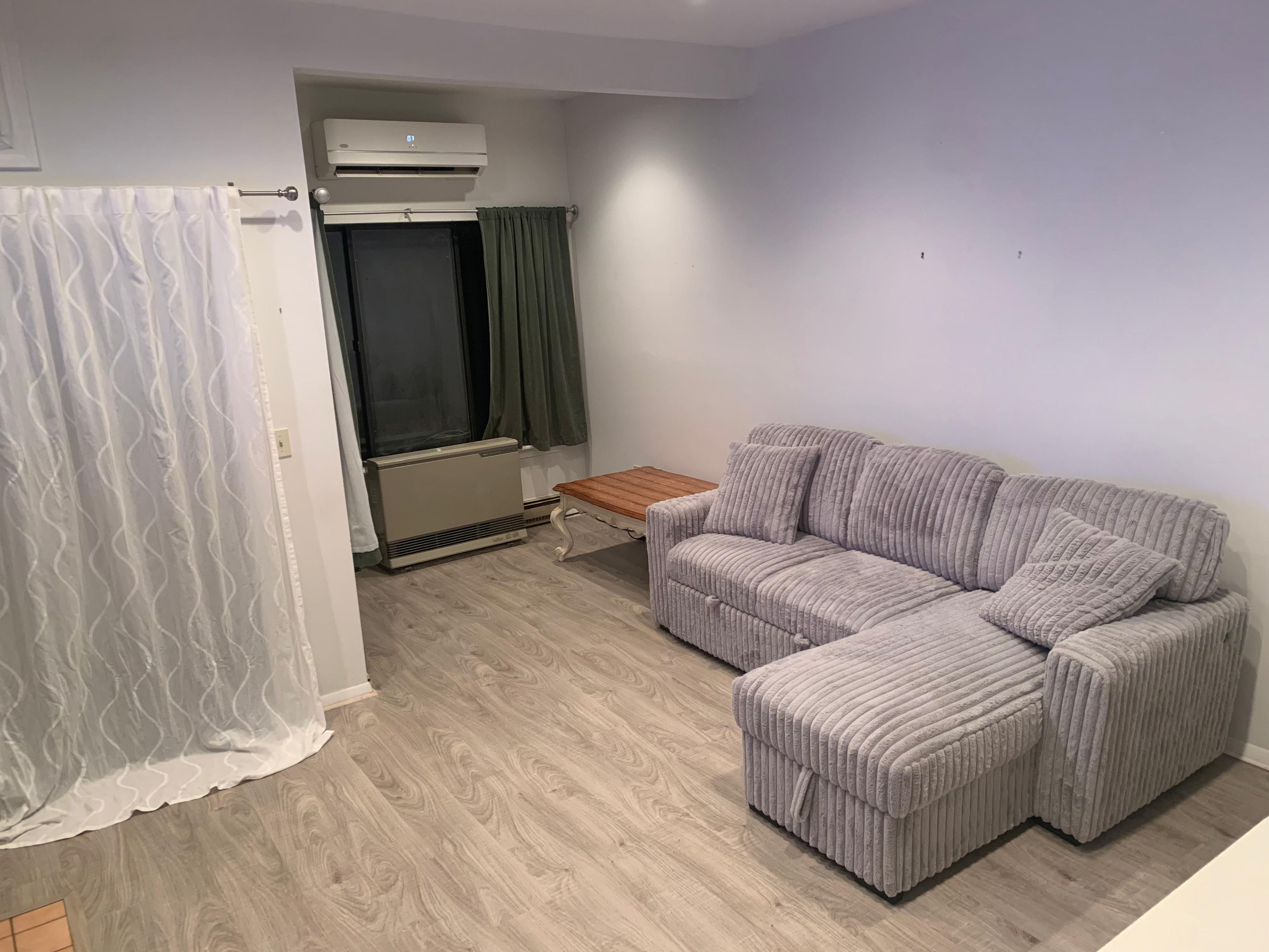 A room with a sectional sofa, a small wooden table, a radiator, and a window with curtains.
