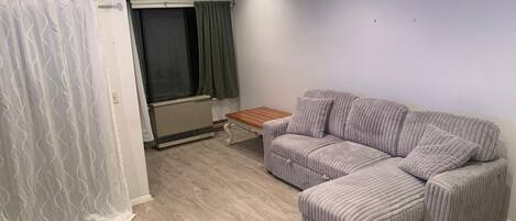 A room with a sectional sofa, a small wooden table, a radiator, and a window with curtains.