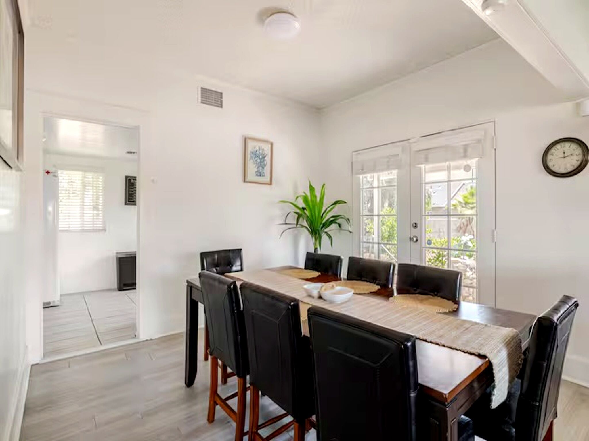 A dining area with a wooden table, black chairs, a potted plant, and a clock on the wall.