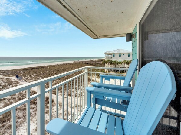 A balcony with a view of the beach and ocean, a blue chair, and a building in the background. - Southern Sunshine, Beachfront Living — Fully Updated & Steps to the Sand (Navarre Beach)