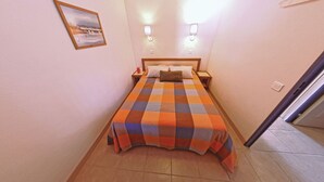 Quarto de hotel pequeno e estreito com colcha xadrez, duas luminárias de cabeceira, um criado-mudo e um quadro na parede.