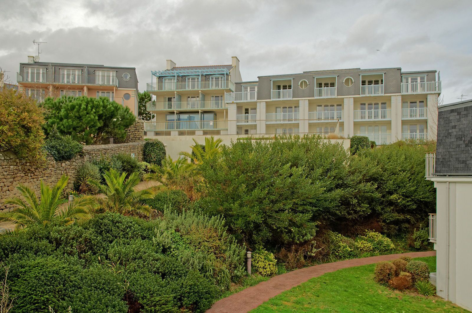 A multi-story residential building with balconies, surrounded by lush greenery and a pathway.