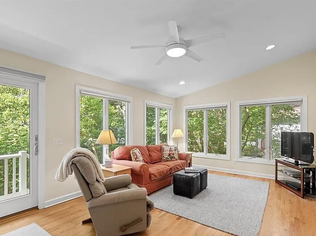 A spacious living room with a ceiling fan, large windows, a sofa, a chair, a TV stand, and a fireplace.