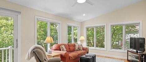 A spacious living room with a ceiling fan, large windows, a sofa, a chair, a TV stand, and a fireplace.