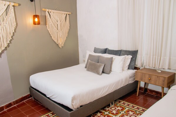 Una cama con estructura de madera, una mesita de noche, adornos de macramé en la pared y una alfombra estampada.