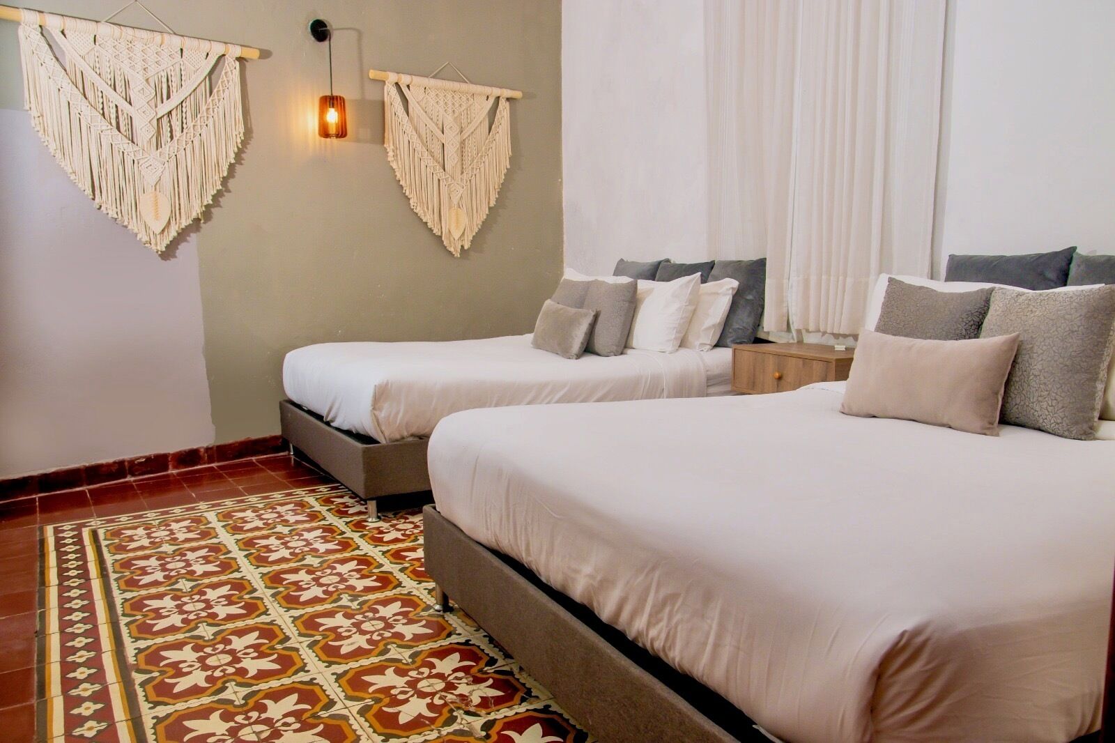 A hotel room with two beds, a patterned rug, macramé wall hangings, and a bedside lamp.
