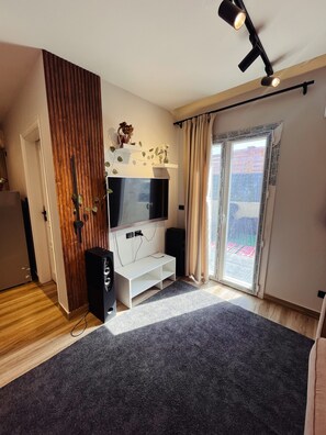 A modern living room with a flat-screen TV, wooden panel wall, and a sliding glass door leading to a balcony.