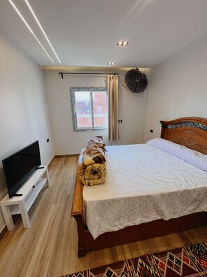 A bedroom with a bed, a TV on a stand, a window with curtains, and a patterned rug.