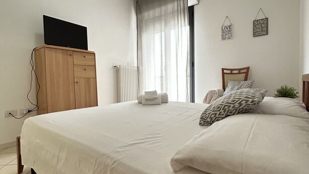 Une chambre à coucher avec un lit, une armoire en bois, un téléviseur posé sur le dessus, un radiateur et une fenêtre avec des rideaux.