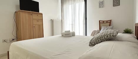 A bedroom with a bed, a wooden wardrobe, a television on top, a radiator, and a window with curtains.