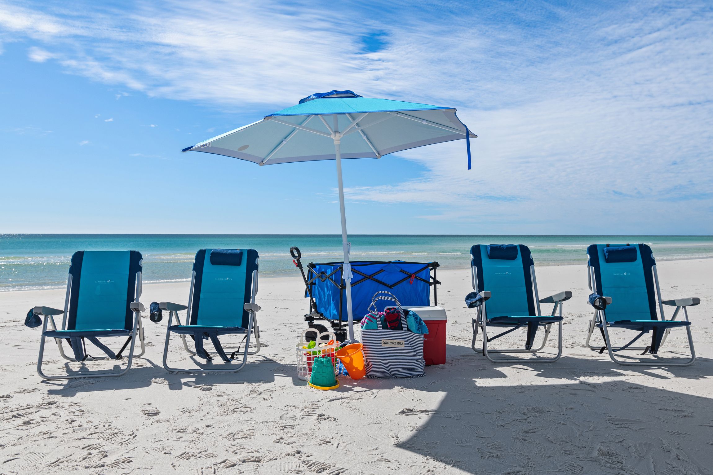 A beach scene with four blue beach chairs, a blue and white umbrella, a cooler, and beach toys.