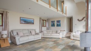 Ein geräumiges Wohnzimmer mit einer großen Eckcouch, einer kleineren Couch und einem Couchtisch. Zwei Fenster mit Vorhängen, ein Gemälde an der Wand und ein Kamin.