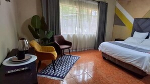 A hotel room with a bed, a coffee table with a coffee pot, a chair, a large plant, and a window with curtains. - Neema Lodge (Boksburg)