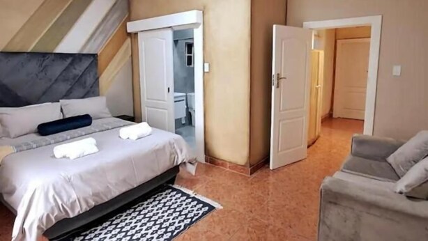 A hotel room with a bed, a chair, a wardrobe, and a door leading to another room. - Neema Lodge (Boksburg)