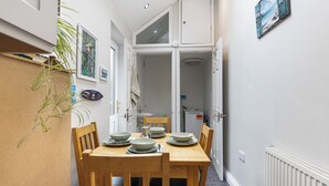 Un couloir étroit mène à une salle à manger où une table est dressée pour deux personnes, avec des chaises en bois, et d’où l’on aperçoit la cuisine.