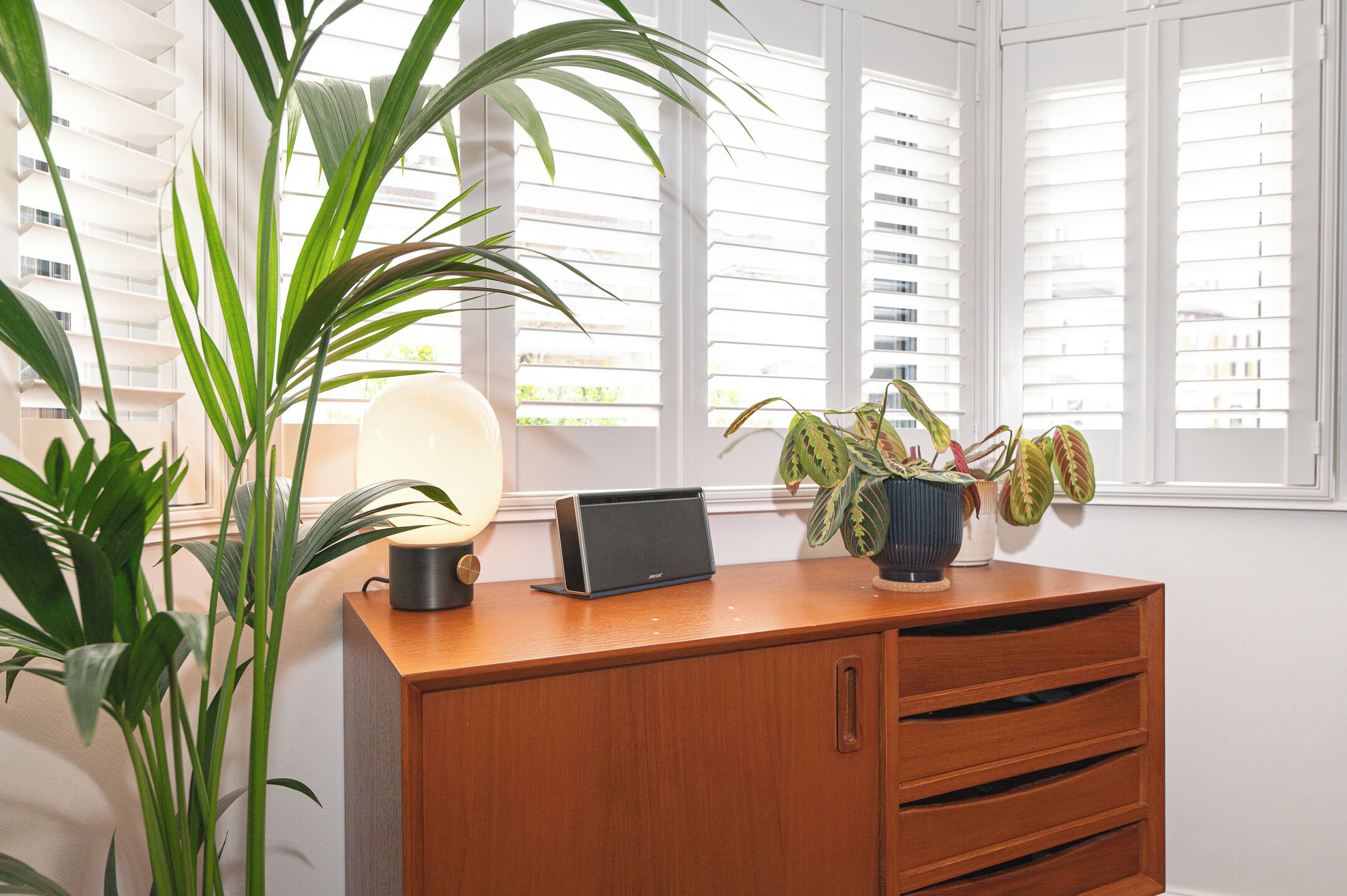 A wooden desk with a laptop, a lamp, and potted plants in a room with white walls and large windows.