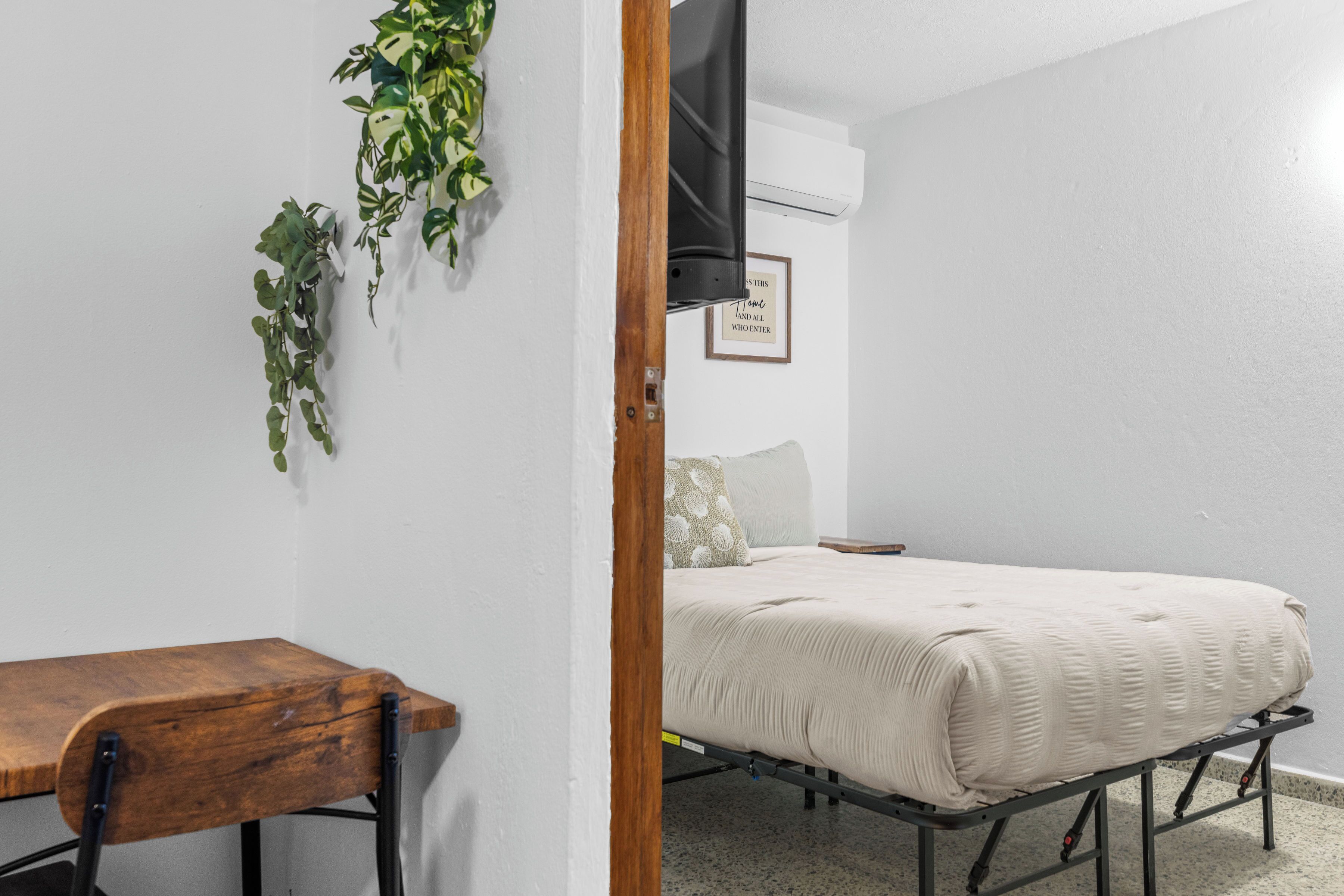 A room with a bed, a wooden desk, a wall-mounted TV, and a framed quote.
