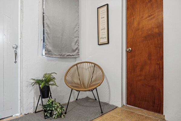 Una puerta de madera, una cita enmarcada en la pared, una silla de mimbre y dos plantas en macetas.