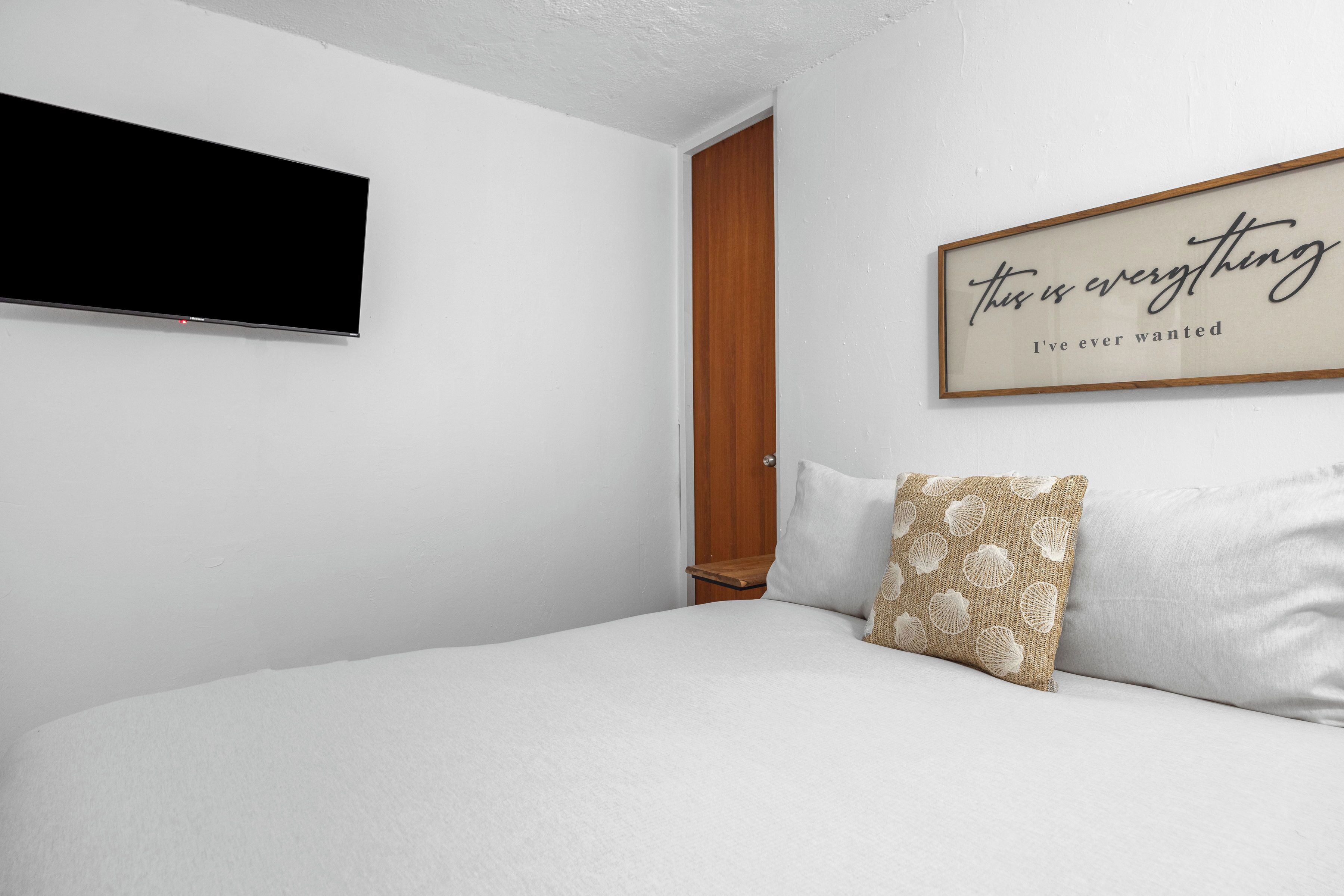 Un dormitorio con una cama, un televisor de pantalla plana montado en la pared, una puerta de madera y una cita inspiradora en la pared.