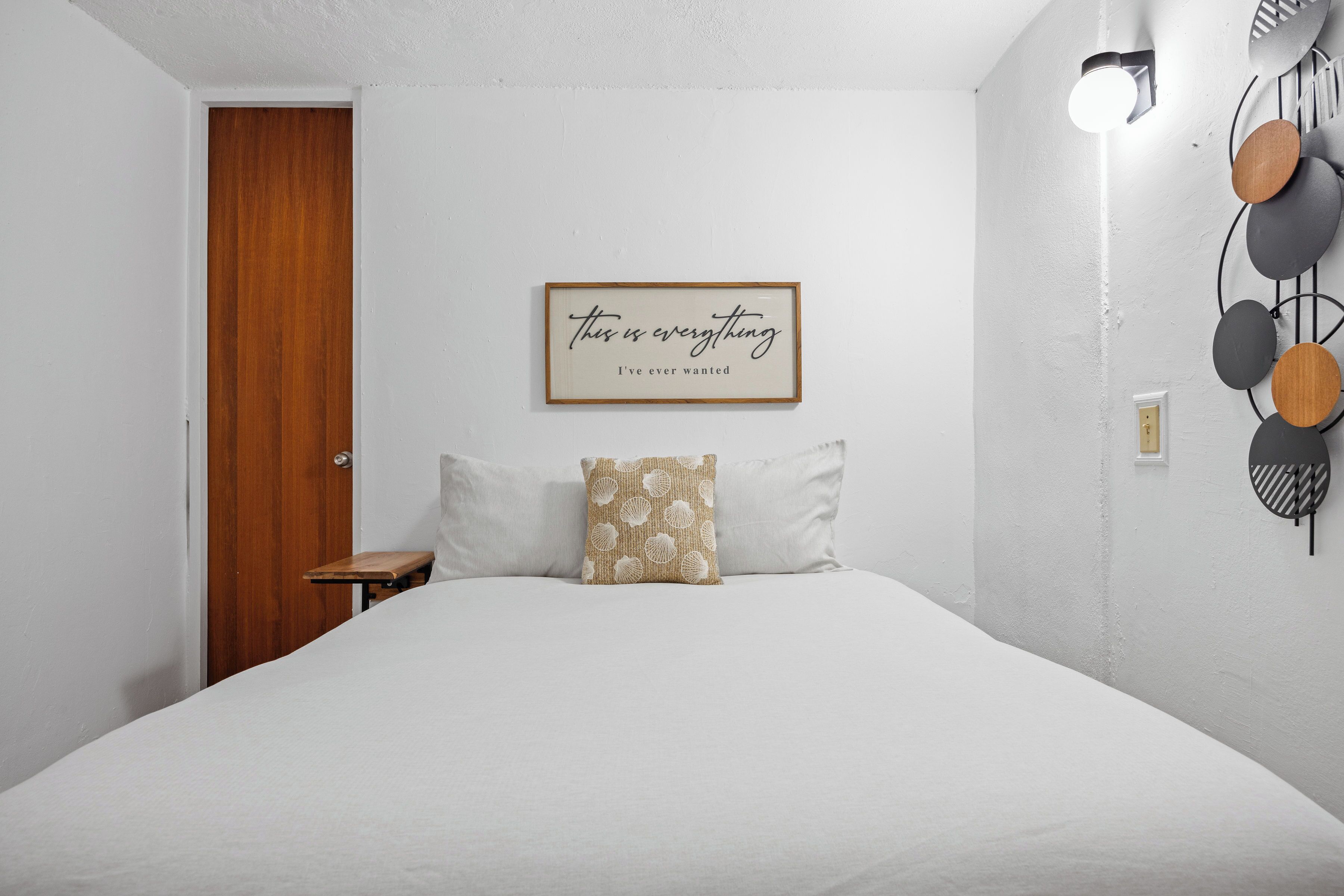 Un dormitorio con puerta de madera, una cita enmarcada en la pared, una cama con ropa de cama blanca y una lámpara fijada a la pared.