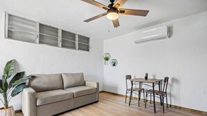 Een kamer met een bank, een eettafel met stoelen, een plafondventilator en een airconditioning.