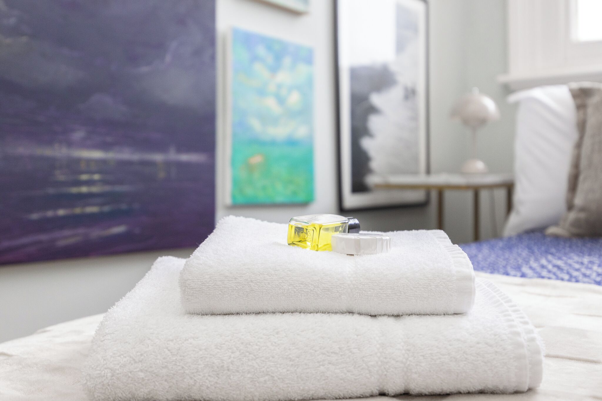 A neatly folded white towel with a bottle of lotion on a bed with a blue blanket.
