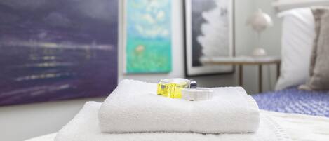 A neatly folded white towel with a bottle of lotion on a bed with a blue blanket.